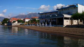 A coastal hotel with a beachside view, including lounge chairs and umbrellas, and a mountain backdrop.