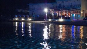 Un hotel frente al agua con piscina que refleja luces, un área exterior cubierta y un edificio de varios niveles.