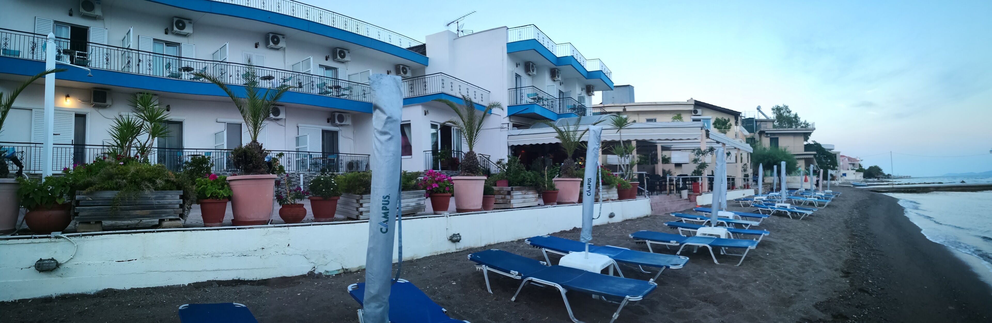Un hotel sulla spiaggia con diversi balconi, piante in vaso e lettini prendisole lungo la costa sabbiosa.