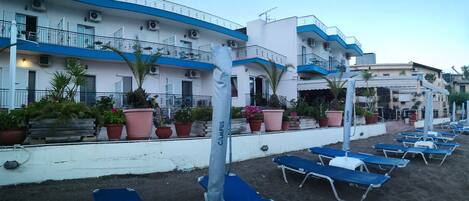 Un hotel sulla spiaggia con diversi balconi, piante in vaso e lettini prendisole lungo la costa sabbiosa.