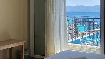 Habitación de hotel con cama, televisión, balcón con piscina y sillas, y vista al mar.