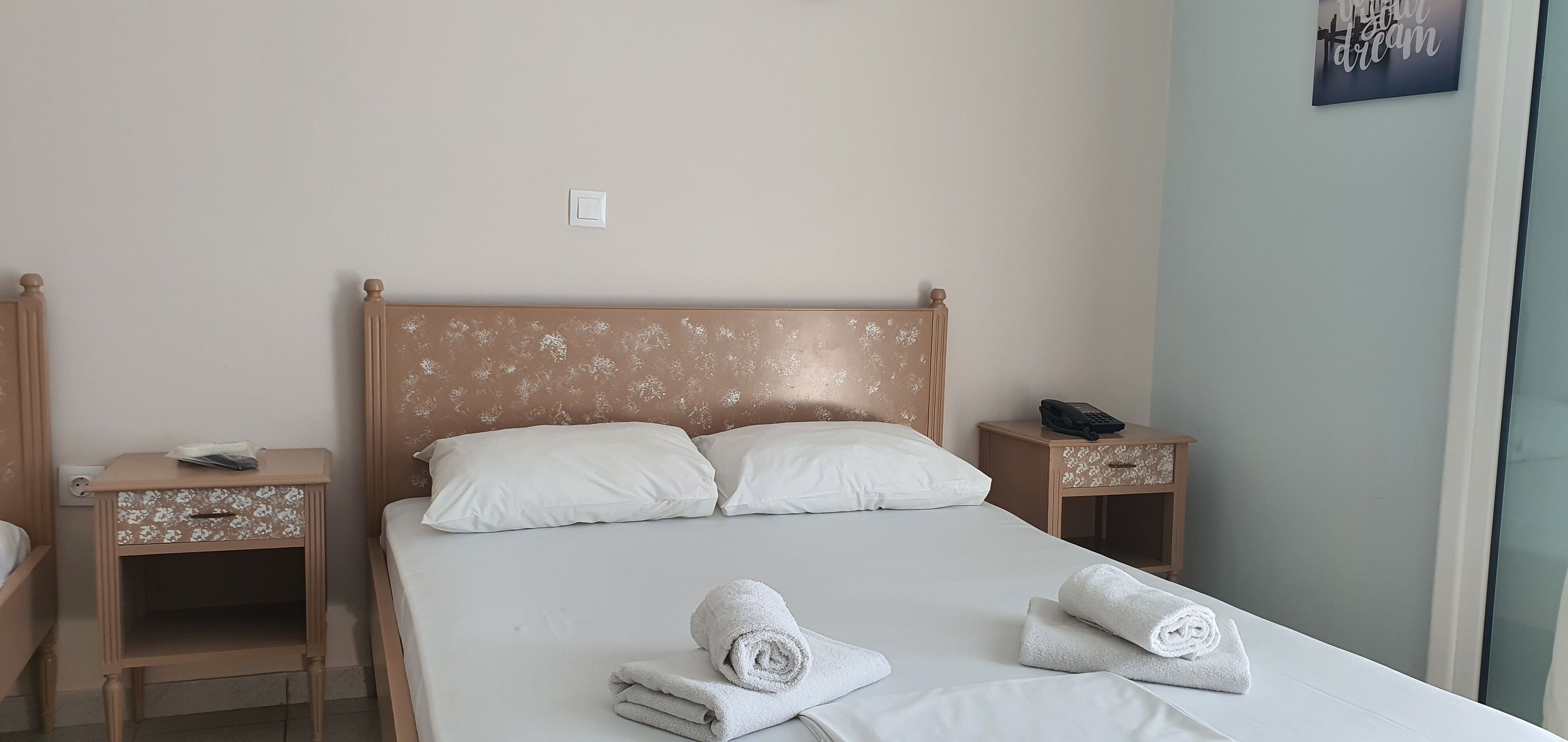 Una cama bien tendida con sábanas blancas y dos toallas enrolladas, una mesita de noche de madera con un teléfono y un objeto decorativo, y un interruptor de luz en la pared.