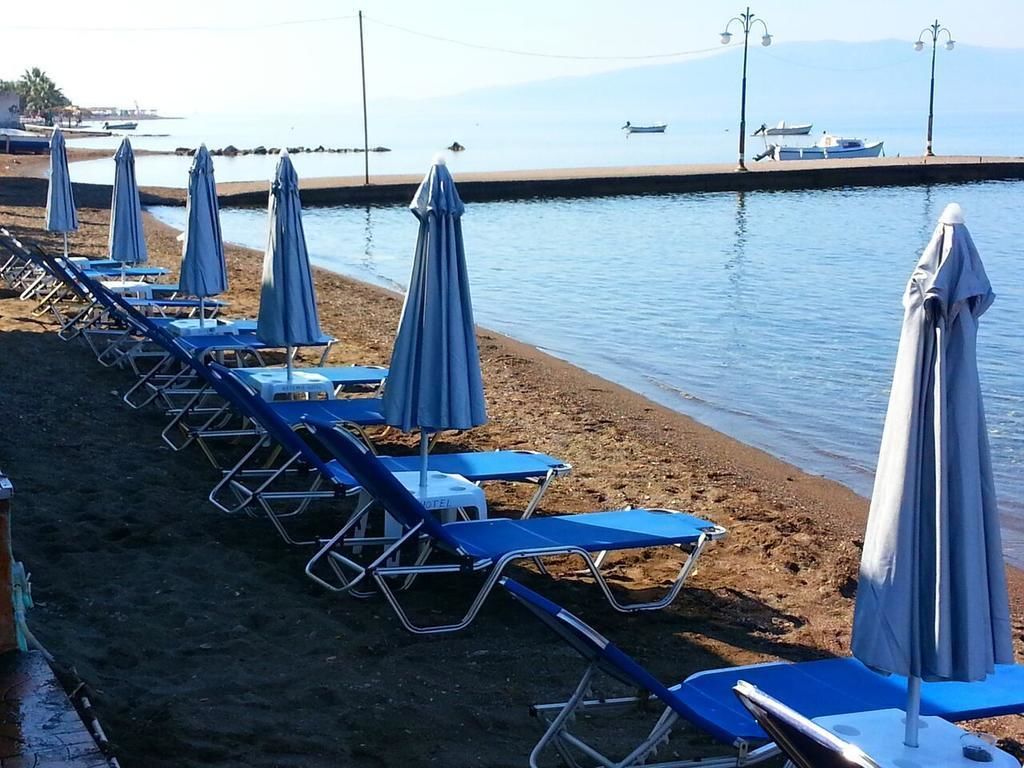 A beach with rows of blue lounge chairs and umbrellas, a sandy shore, and a calm sea with boats in the background.
