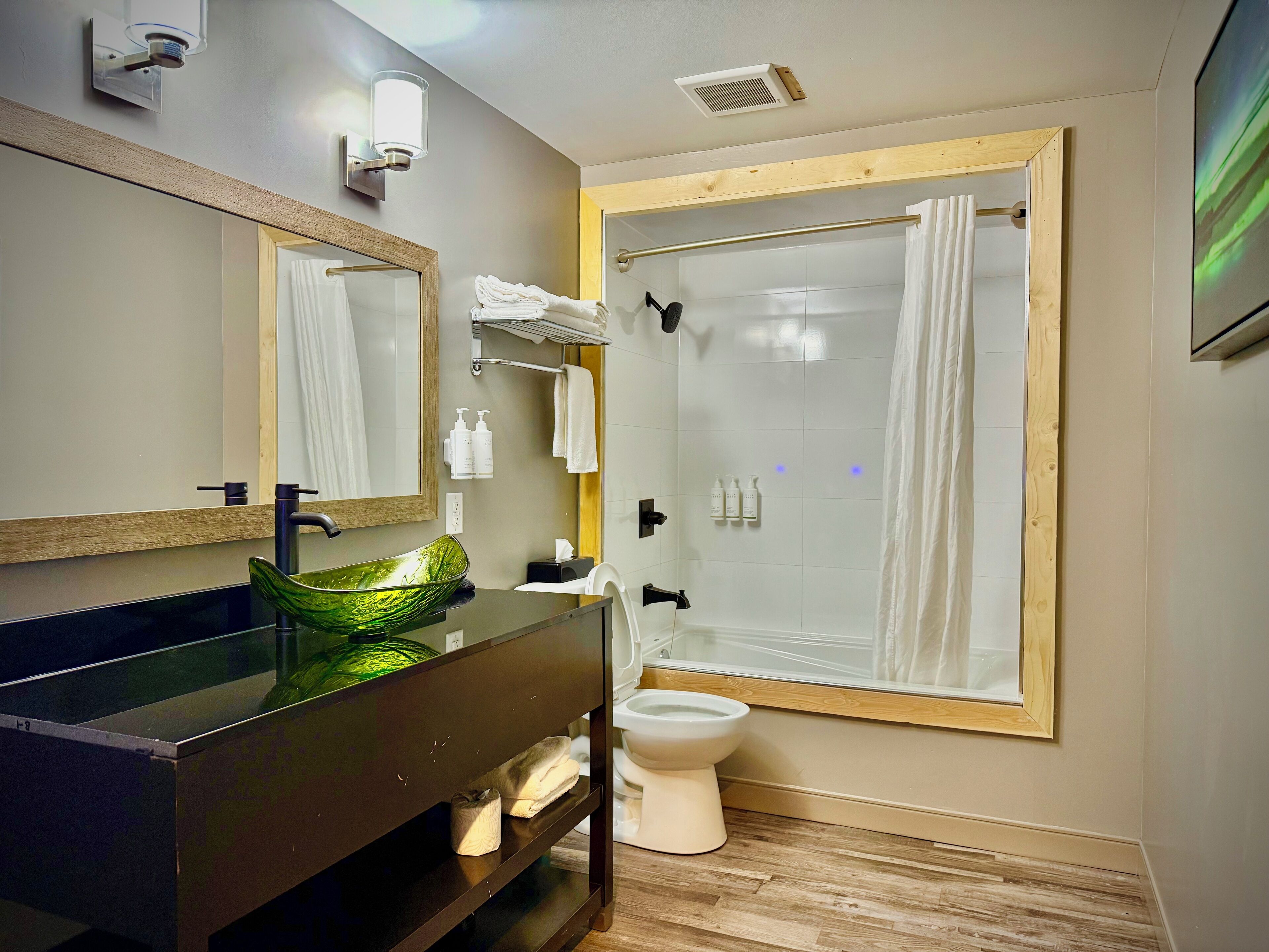 A modern bathroom with a large mirror, a glass bowl sink, a toilet, and a shower with a wooden frame.