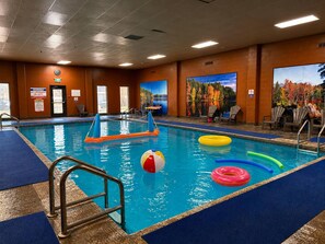 An indoor swimming pool with a lifeguard station, poolside chairs, and a mural of autumn foliage.