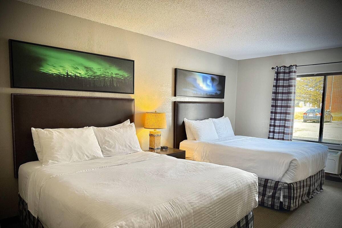 Habitación de hotel con dos camas, cabecera, lámpara, un cuadro y ventana con cortinas.