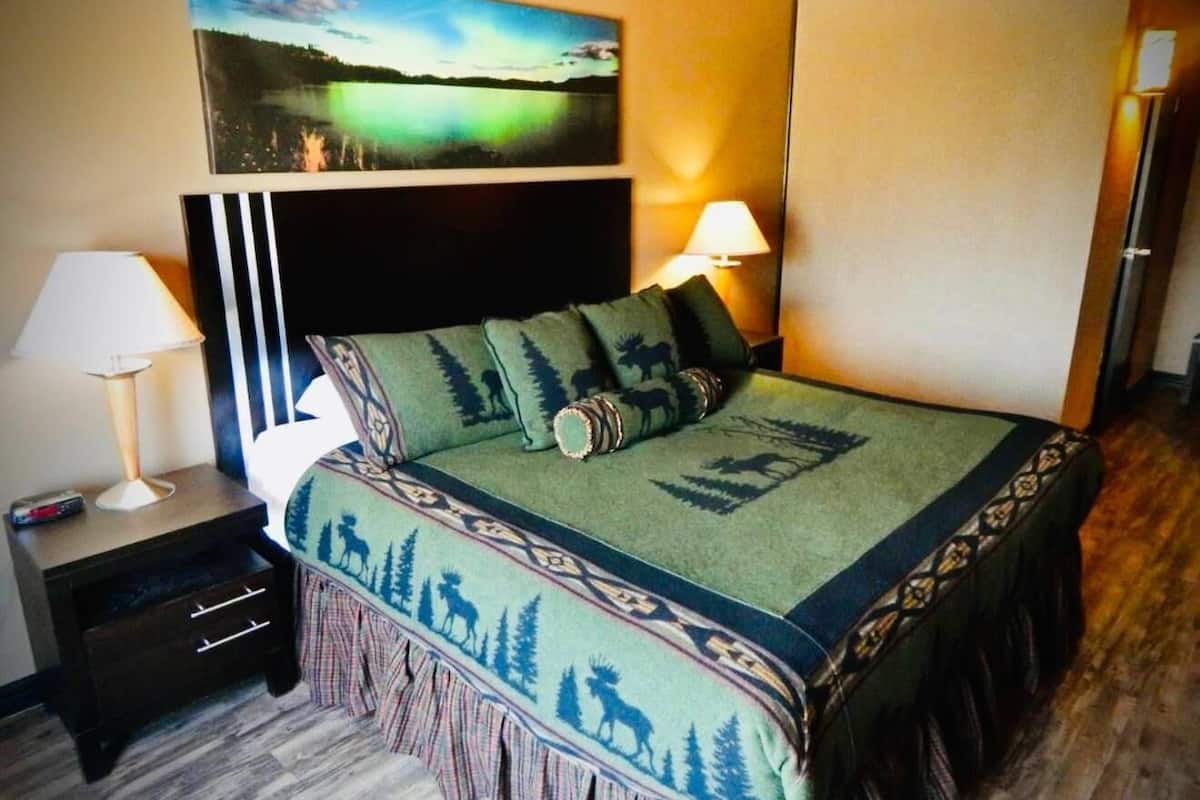 Una cama con una manta y almohadas verdes, una mesita de noche con una lámpara, un cuadro en la pared y un piso de madera.