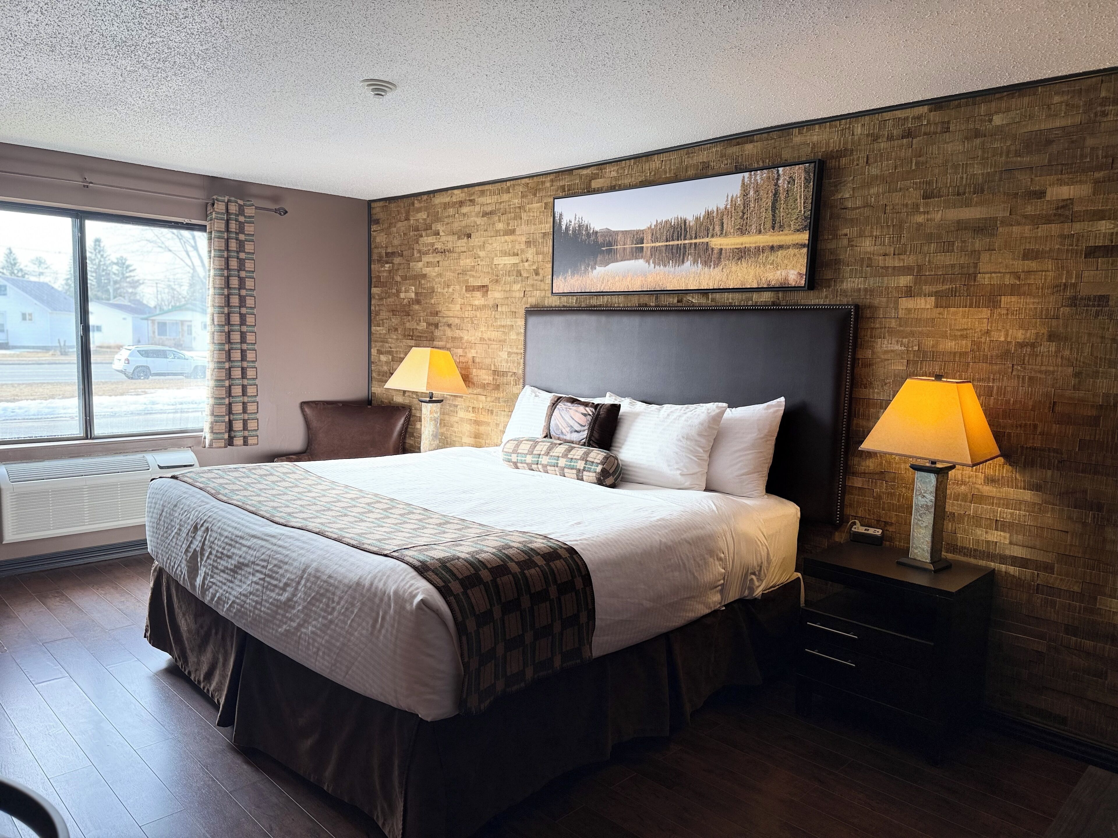 A hotel room with a large bed, two bedside lamps, a headboard with a painting, a window with a view of houses, and a wooden floor.