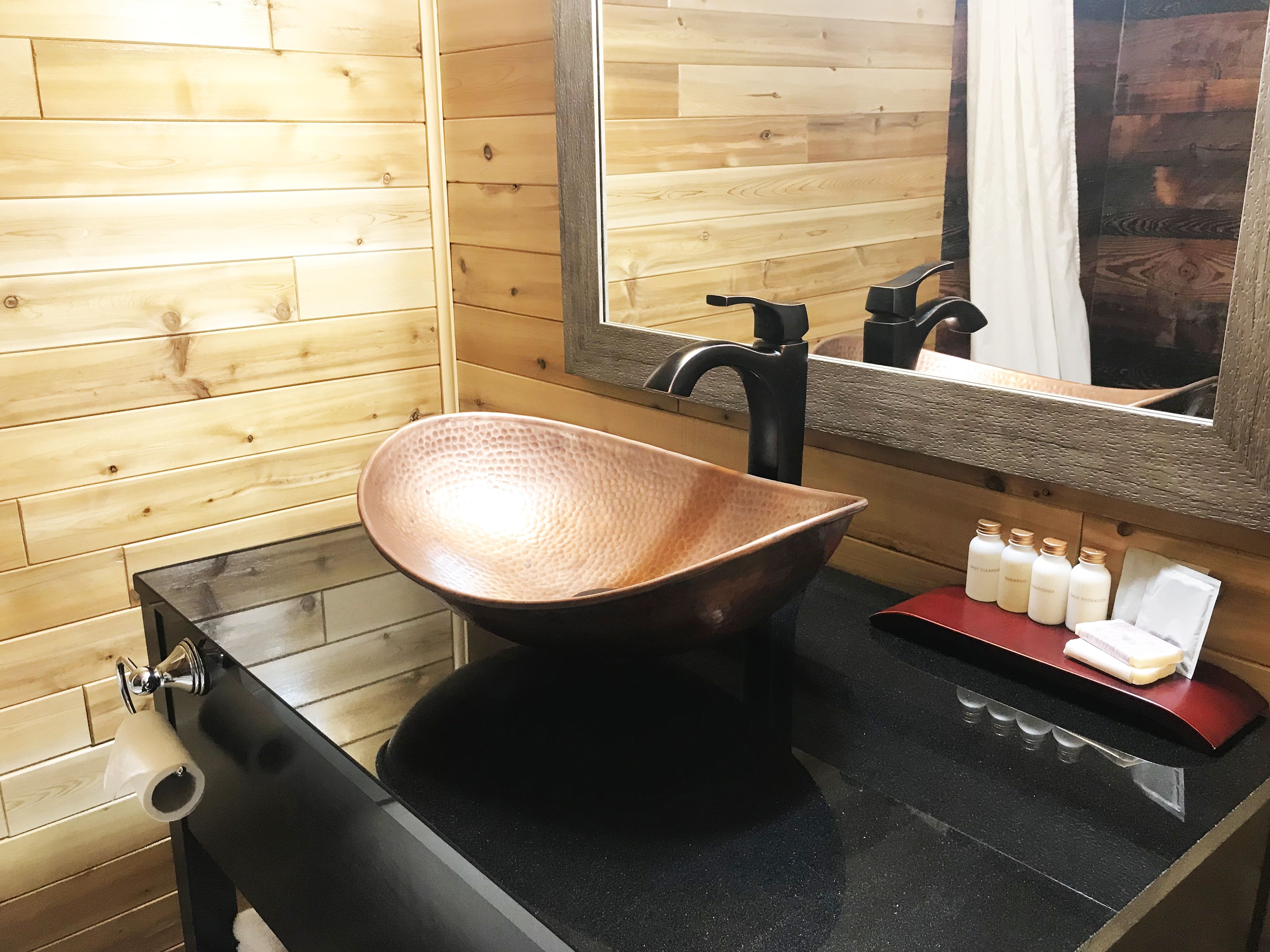 A wooden wall, a black sink with a copper bowl, a mirror, and toiletries on a counter.