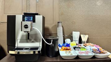 A coffee machine with a digital display, a trash bin, and a buffet with various food items and utensils.