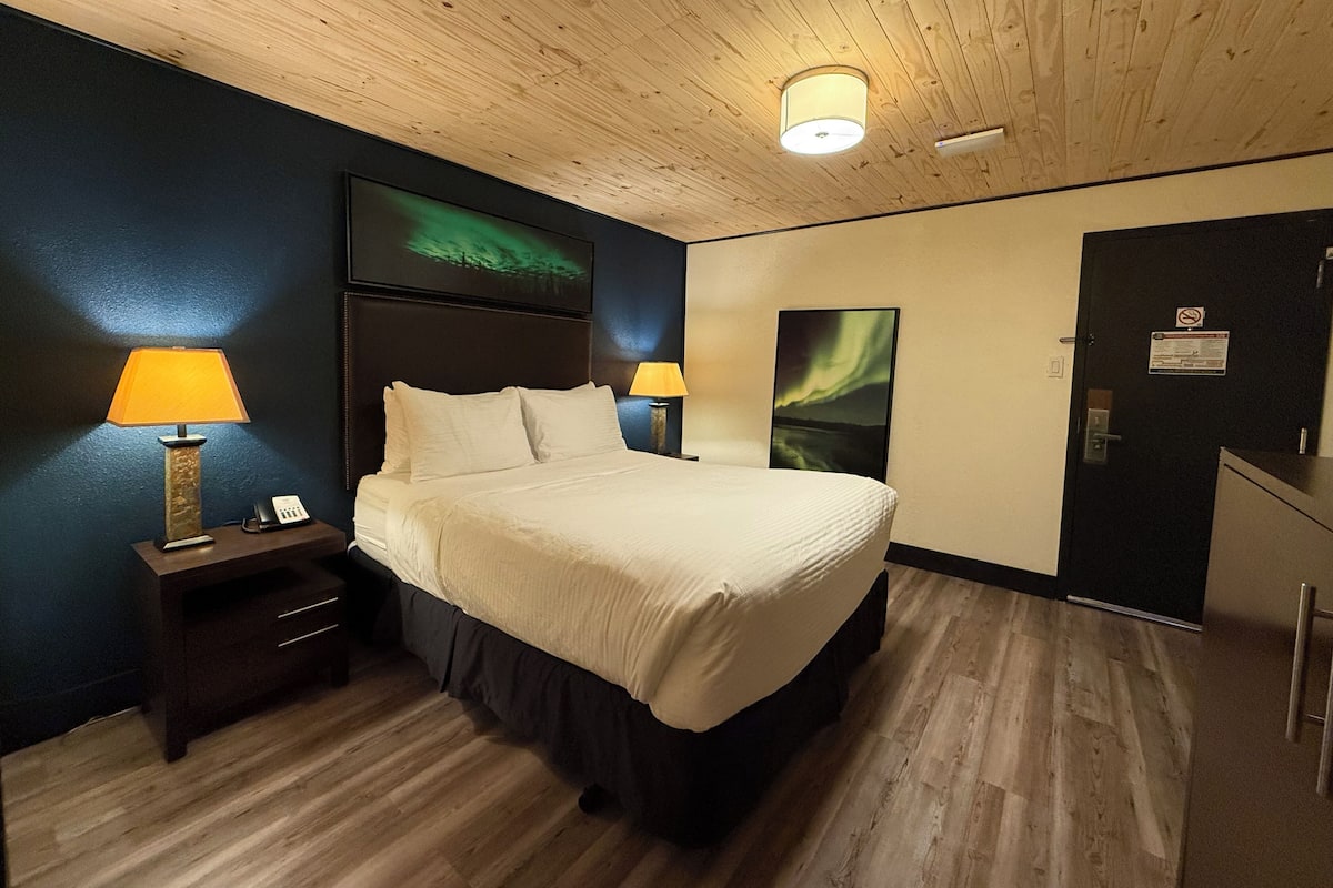 Habitación de hotel con techo de madera, cama con ropa blanca, dos mesitas de noche con lámparas y una puerta con letrero.