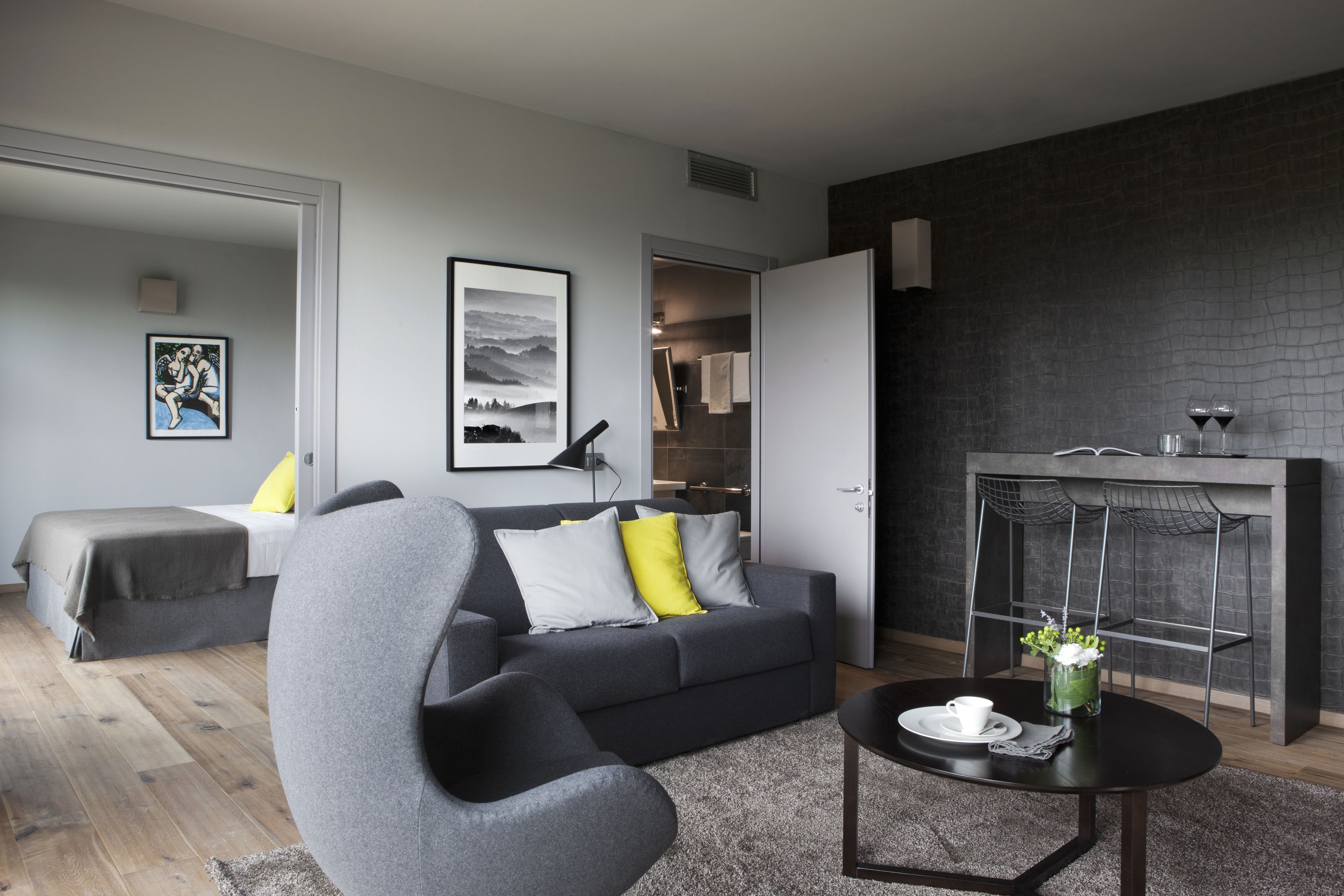 Un salon moderne avec un canapé gris, un fauteuil noir et une table basse. On y trouve également un coin repas avec une chaise en métal et un vase de fleurs.