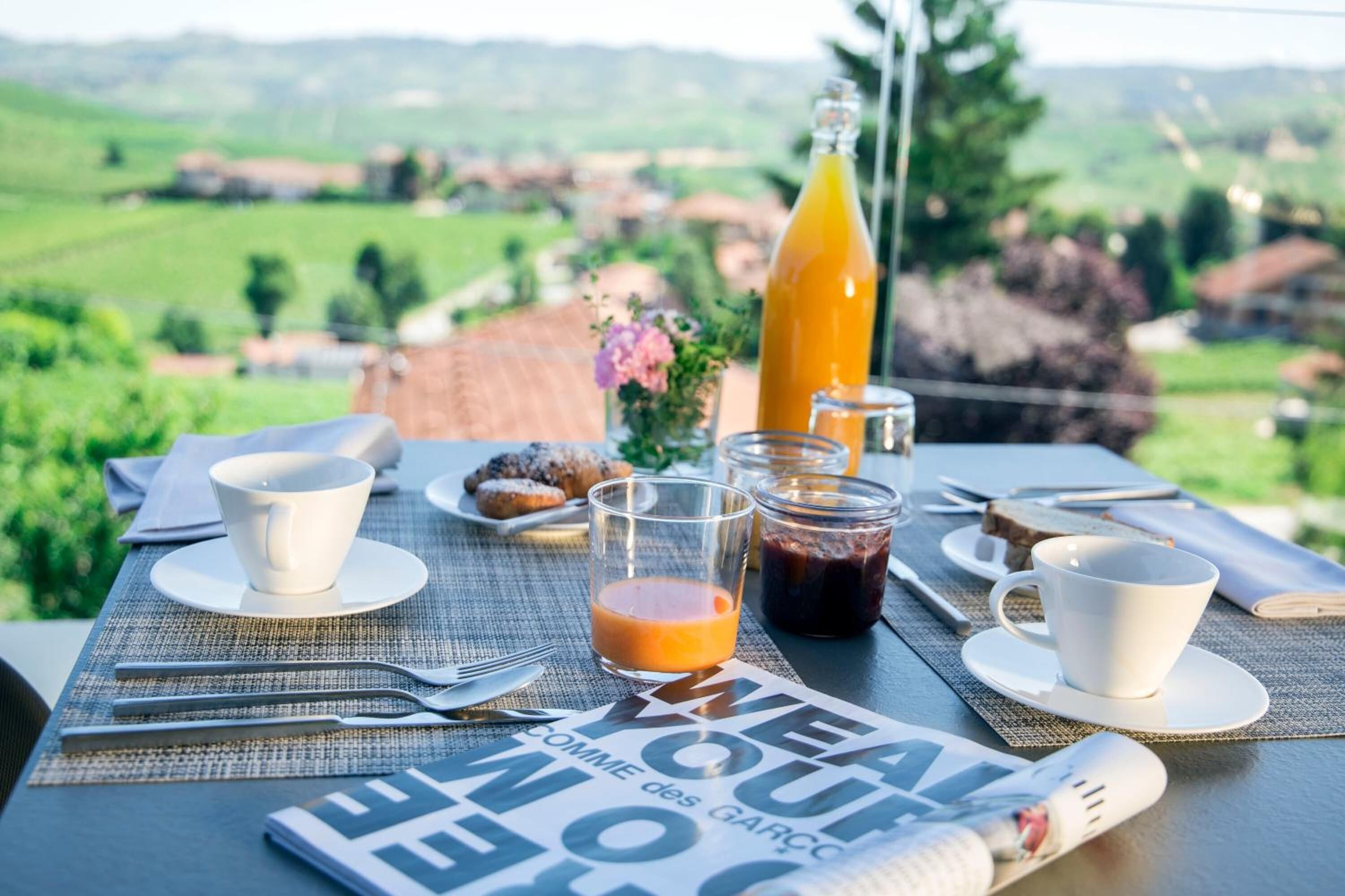 A breakfast table with a view of the countryside, a bottle of orange juice, a glass of juice, a cup, a muffin, and a newspaper.