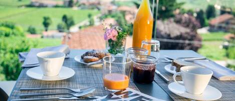 A breakfast table with a view of the countryside, a bottle of orange juice, a glass of juice, a cup, a muffin, and a newspaper.