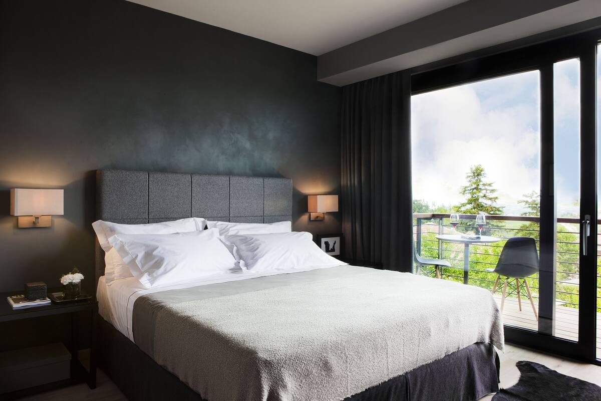 A modern bedroom with a large bed, two bedside tables with lamps, a view of the outdoors through a sliding glass door, and a small balcony area.