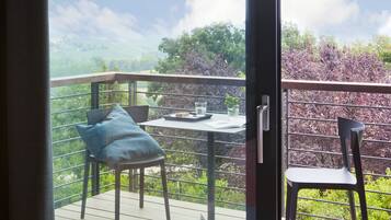 A balcony with a table and chairs, a bed with a red blanket, and a view of greenery and hills.