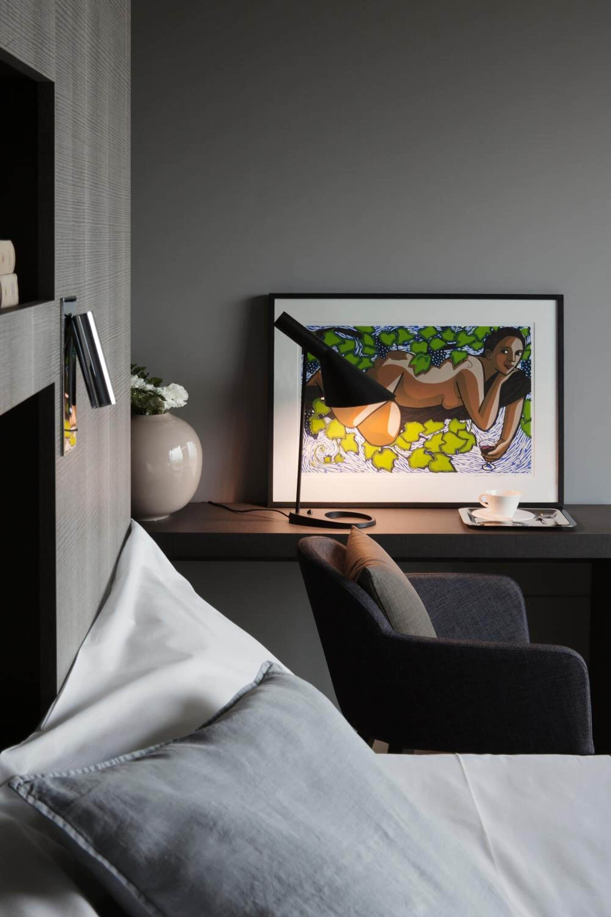 A modern room with a bed, a desk with a framed artwork, a vase with flowers, and a lamp.