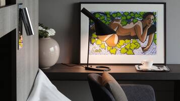 A modern room with a bed, a desk with a framed artwork, a vase with flowers, and a lamp.