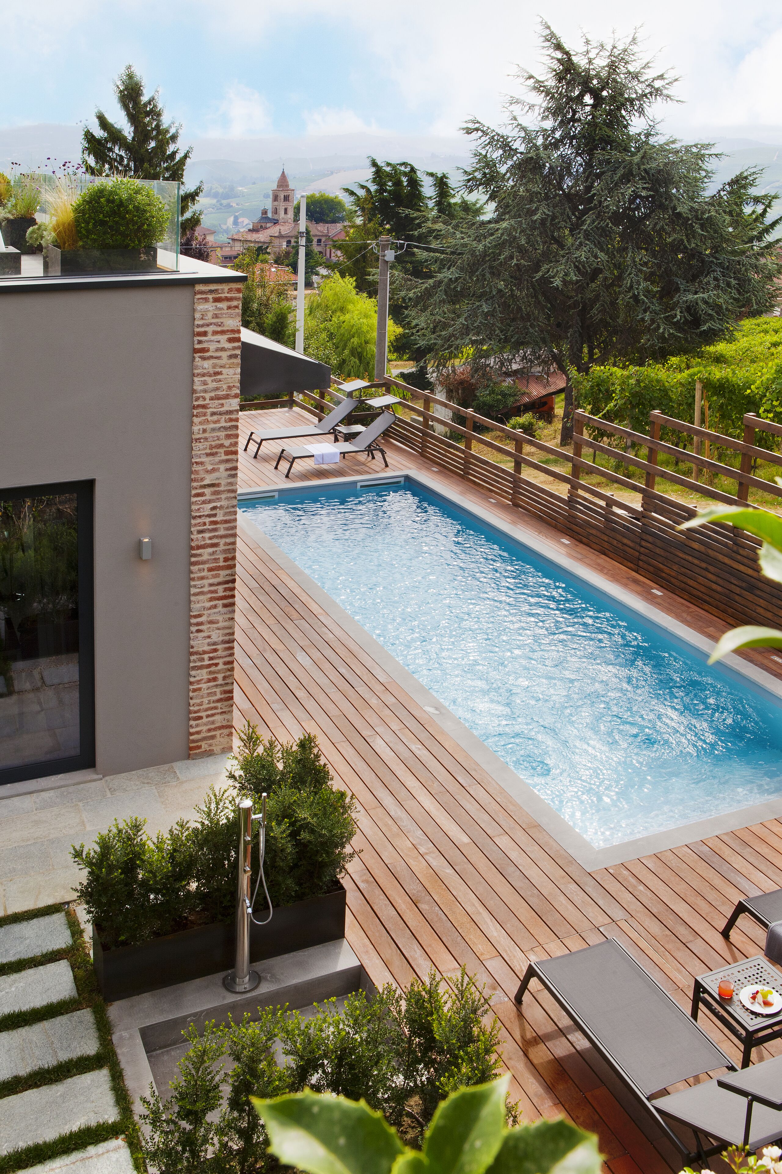 Un área de piscina moderna al aire libre con cubierta de madera, sillas de descanso y vista a un edificio lejano.