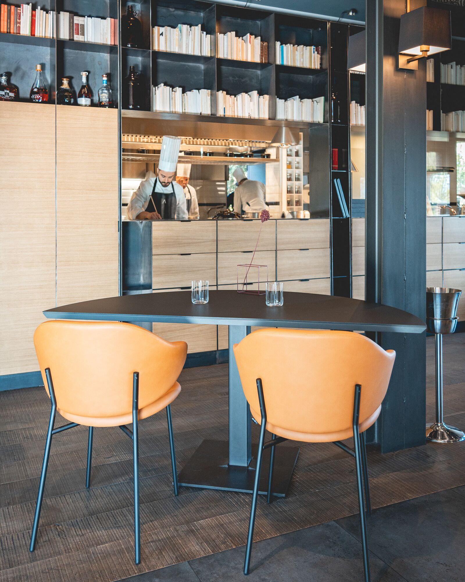 A modern dining area with a round table, two orange chairs, and a bookshelf filled with books and bottles in the background.