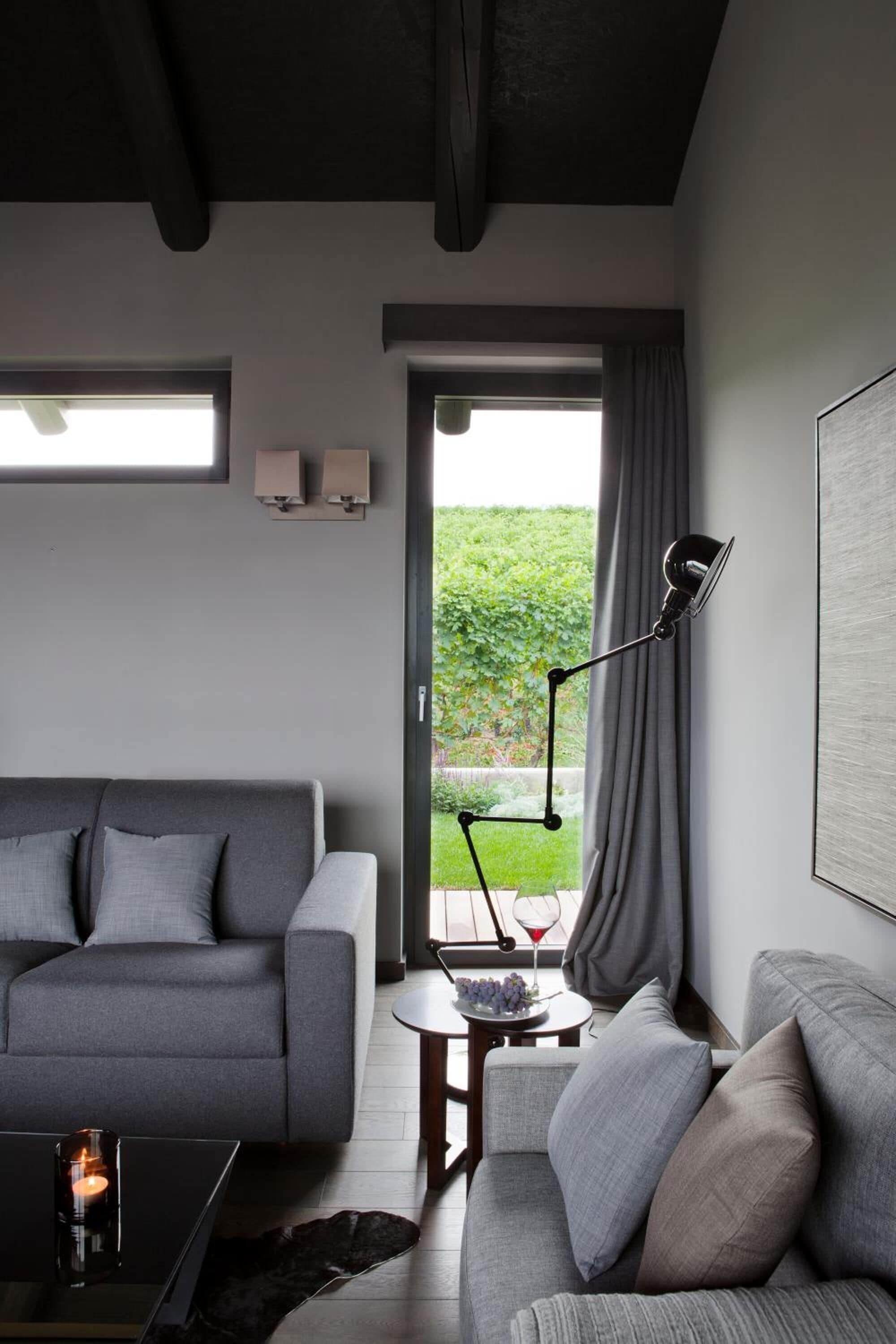 Ein modernes Wohnzimmer mit einem grauen Sofa, einem Couchtisch mit Kerze und einer Stehlampe. Ein Fenster mit Vorhängen bietet Blick auf Grünflächen.
