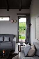 Una sala moderna con un sofá gris, una mesita de centro con una vela y una lámpara de pie. Se observa una ventana con cortinas y vistas a zonas verdes al exterior.