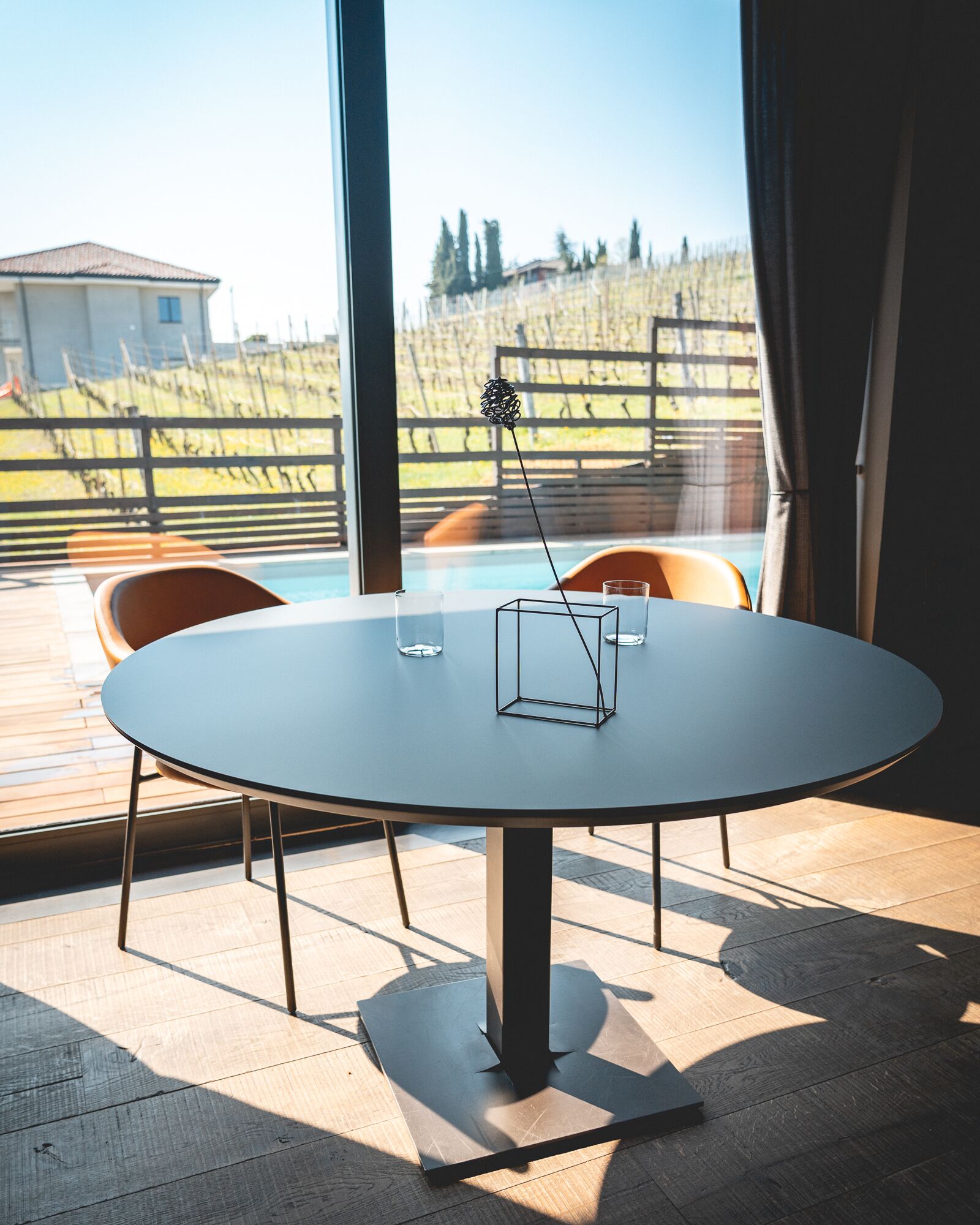 A round table with a metal base and four chairs, a glass on the table, and a view of a vineyard through large windows.