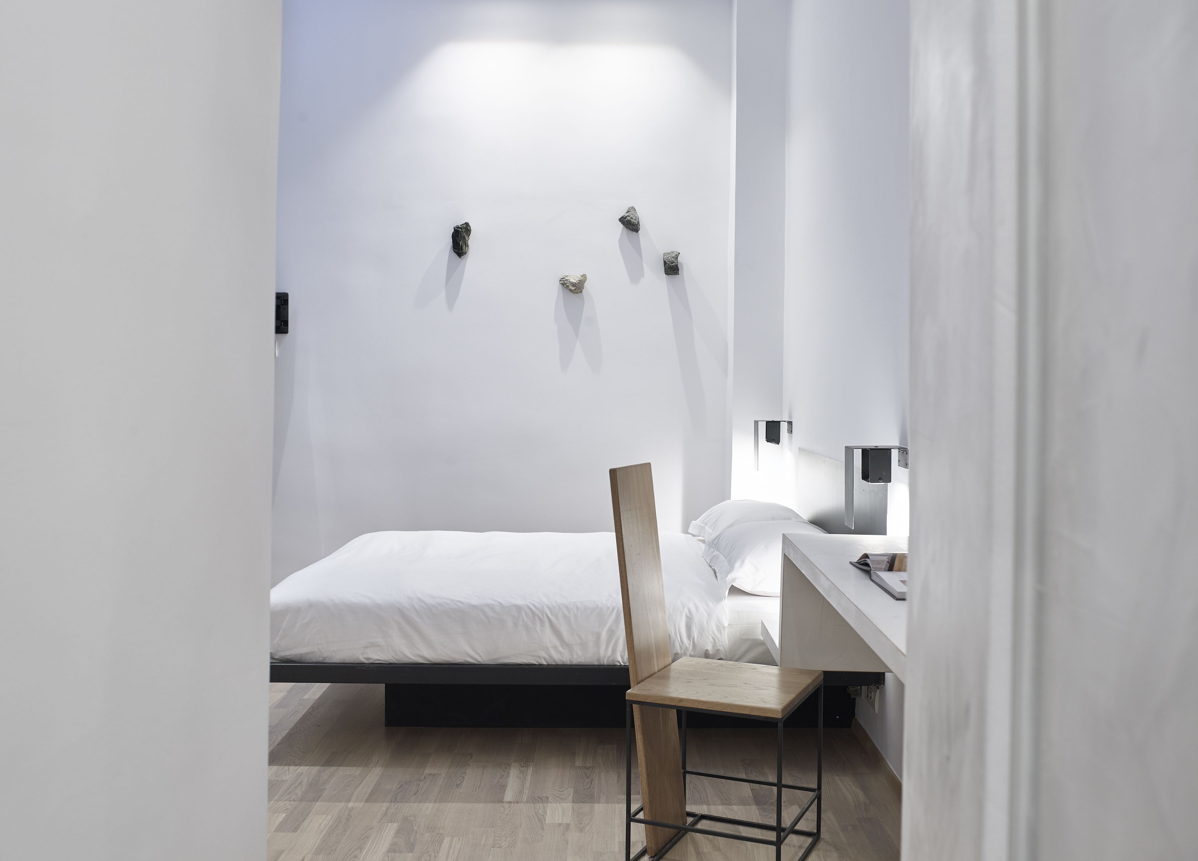 A minimalist hotel room with a bed, a wooden chair, a desk, and wall-mounted lights.