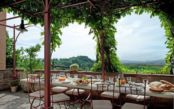 An outdoor dining area with a table set for four, surrounded by a pergola with climbing plants, overlooking a scenic landscape.