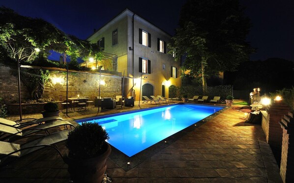 A swimming pool with lounge chairs and a covered patio area, surrounded by a stone wall and trees.
