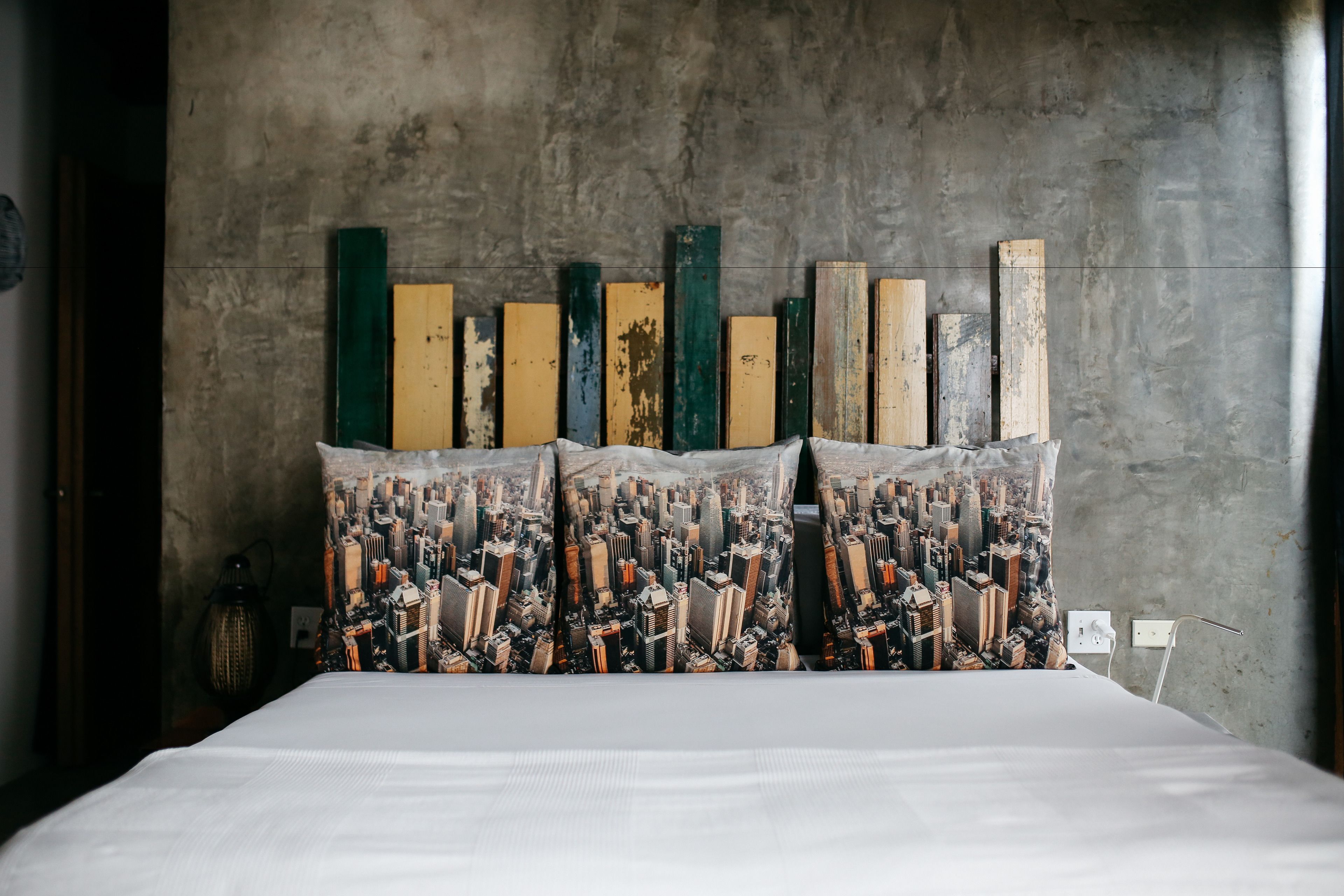 A bed with a cityscape print headboard and pillows, against a concrete wall.