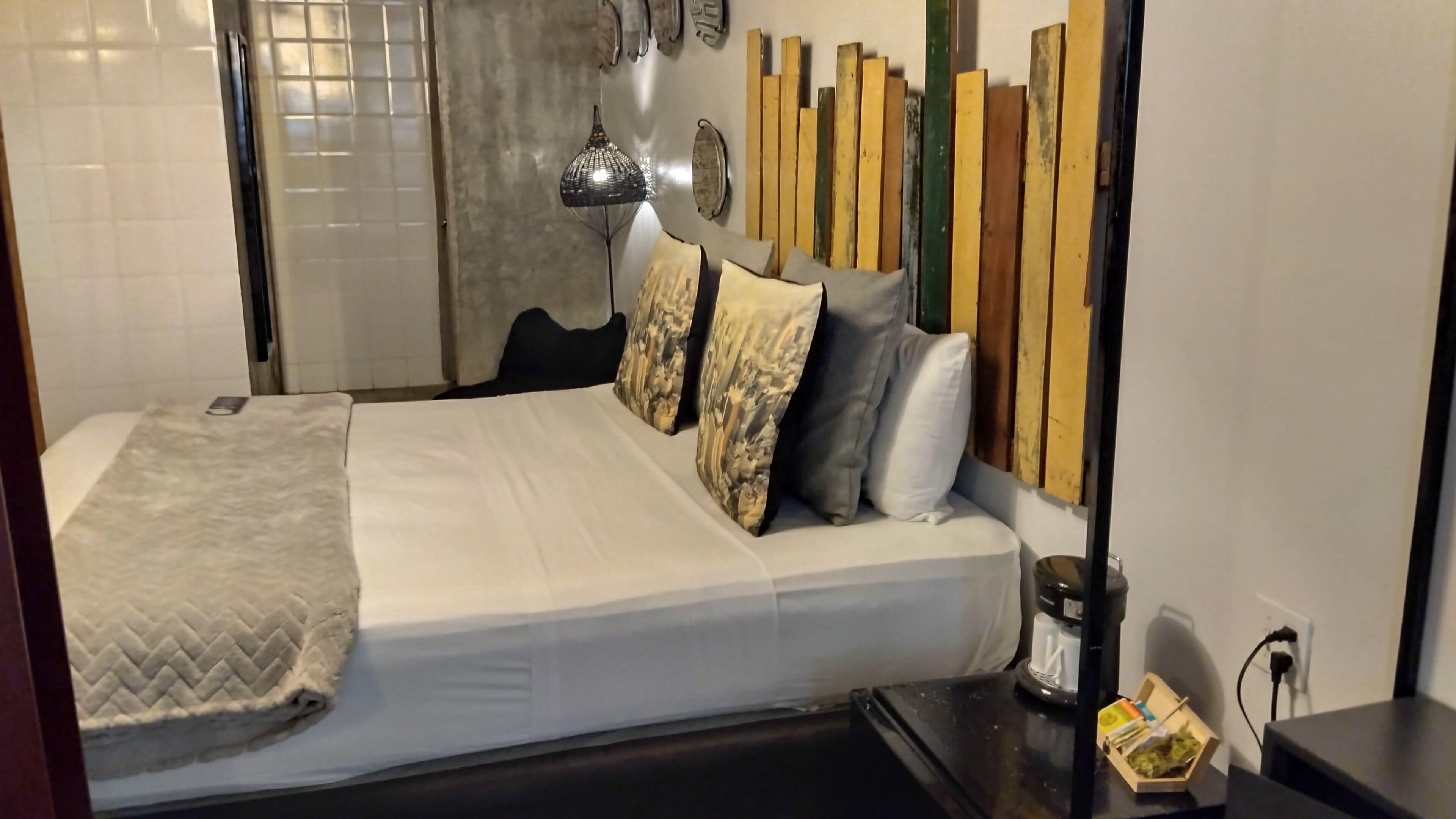 A neatly made bed with a white duvet and patterned pillows, a bedside table with a coffee maker, and a wall with a mix of wooden planks and a decorative metal piece.