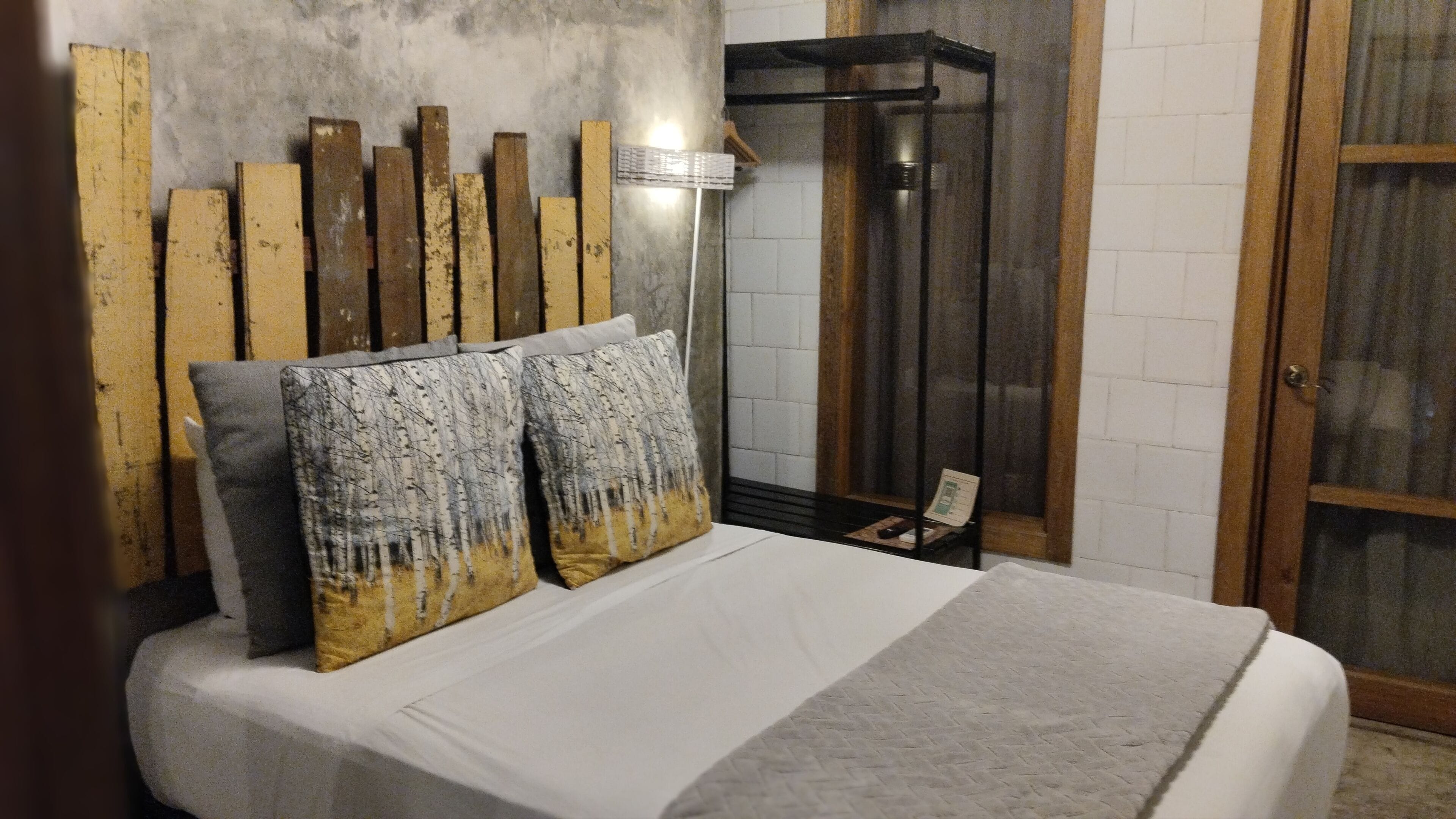 A bed with white bedding and two decorative pillows, a metal shelf unit, and a wooden headboard against a white tiled wall.