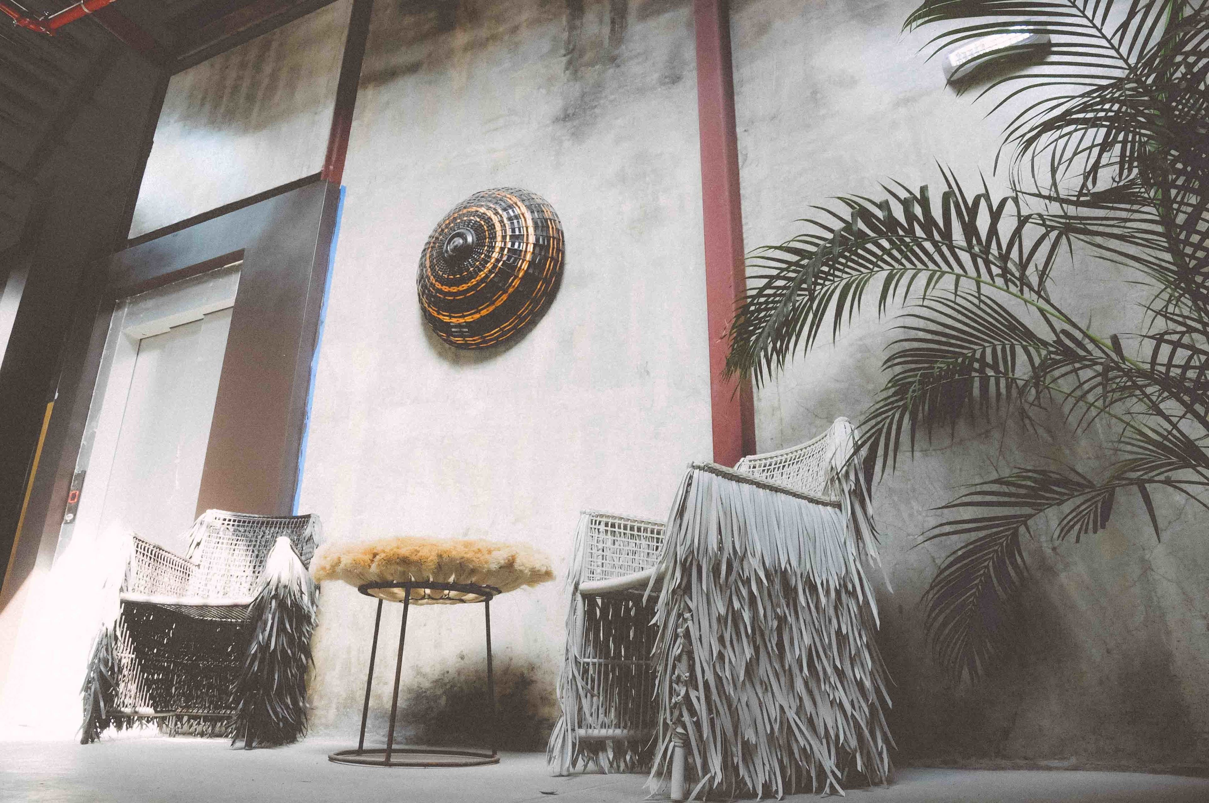 A room with a concrete wall, a woven chair, a small round table, and a decorative wall hanging.