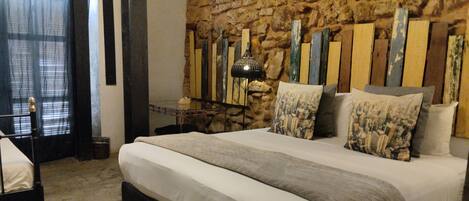 A bedroom with a stone wall, a bed with a grey blanket, and a nightstand with a lamp.
