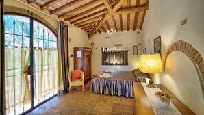 Un dormitori amb sostre de fusta, un llit amb mantell estriat, una cadira, una taula amb una làmpada i una finestra amb cortines.