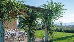 An outdoor dining area with a pergola, tables set for a meal, and a grassy landscape with distant hills. - Villa Danilo (San Casciano dei Bagni)