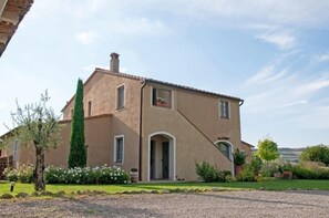 Une maison à deux étages, dotée d’une façade beige, d’une cheminée et d’un jardin bien entretenu où poussent diverses plantes et fleurs.