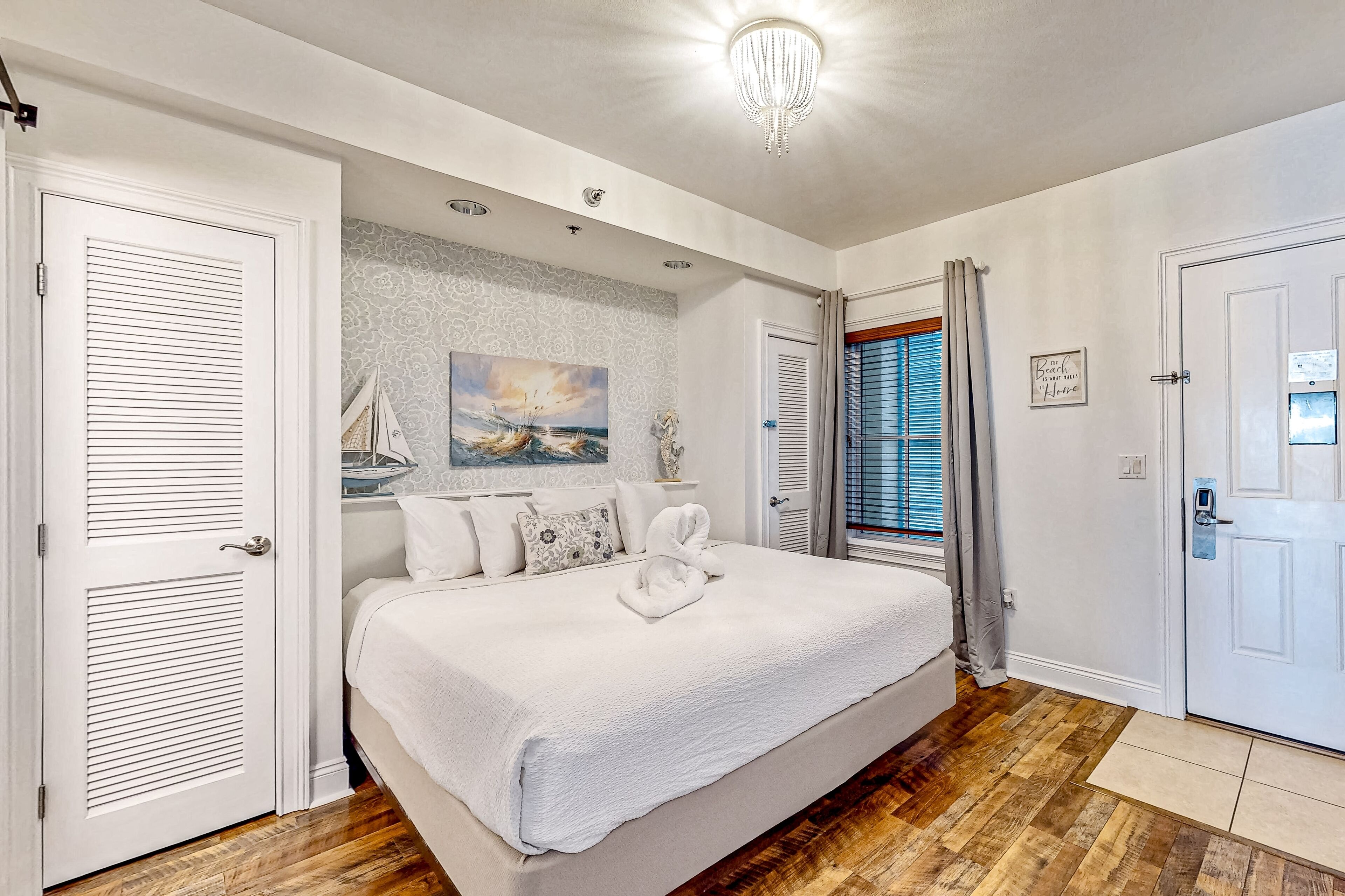 A bedroom with a large bed, white curtains, a chandelier, and a painting on the wall.