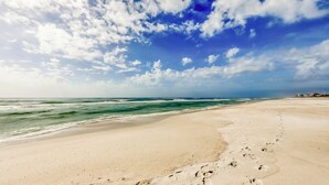 Une vaste plage de sable où l’on peut voir des empreintes de pas menant à l’eau, sous un ciel partiellement nuageux.