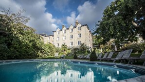A swimming pool with lounge chairs, surrounded by lush greenery and a large building in the background. - The Royal Hotel (Ventnor)