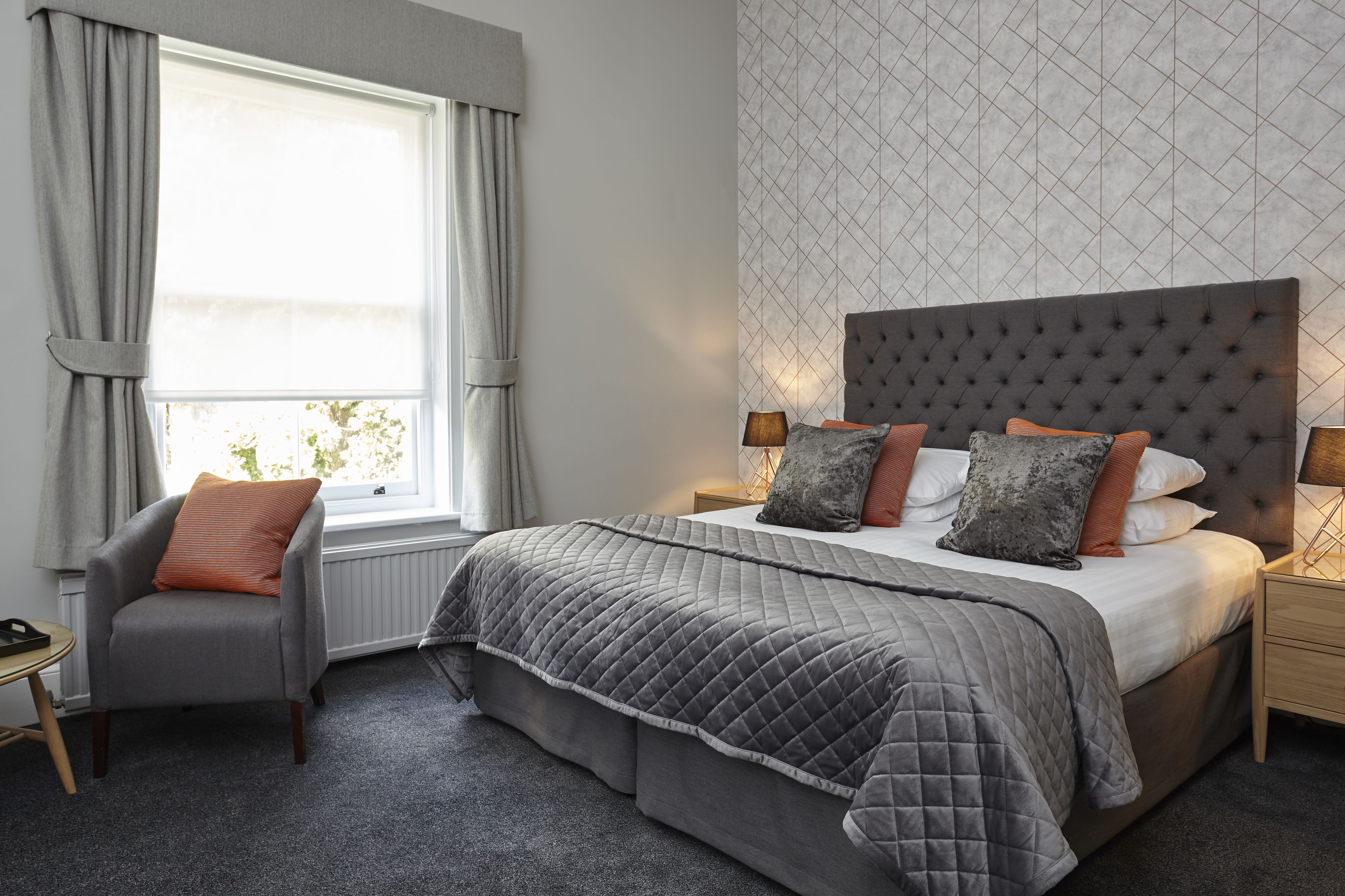 A bedroom with a large bed, grey bedding, a grey armchair, and a window with curtains.