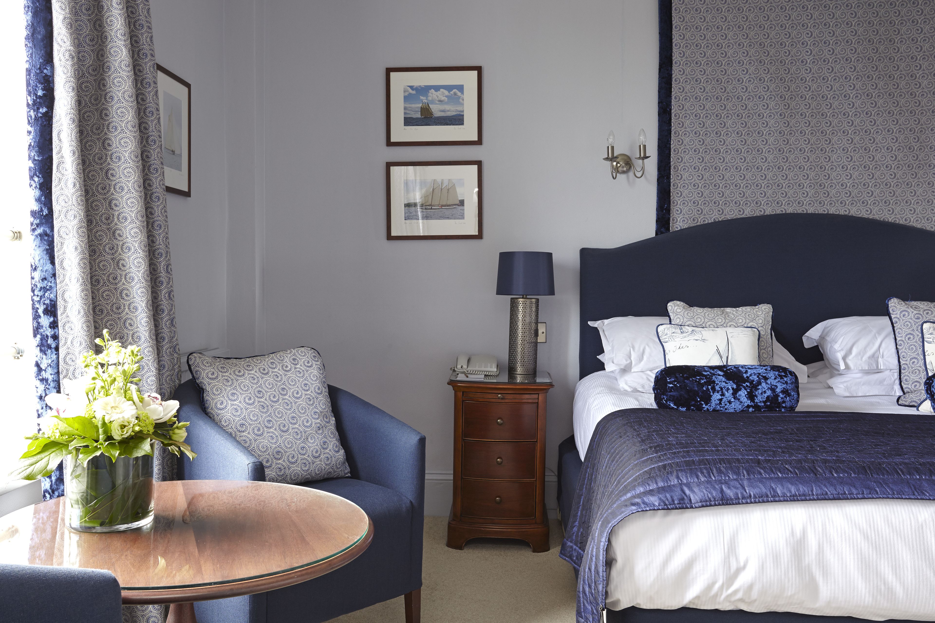 A hotel room with a bed, a bedside table with a lamp, a chair, a small table with a vase of flowers, and two framed pictures on the wall.