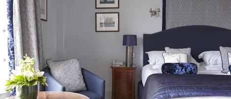 A hotel room with a bed, a bedside table with a lamp, a chair, a small table with a vase of flowers, and two framed pictures on the wall.
