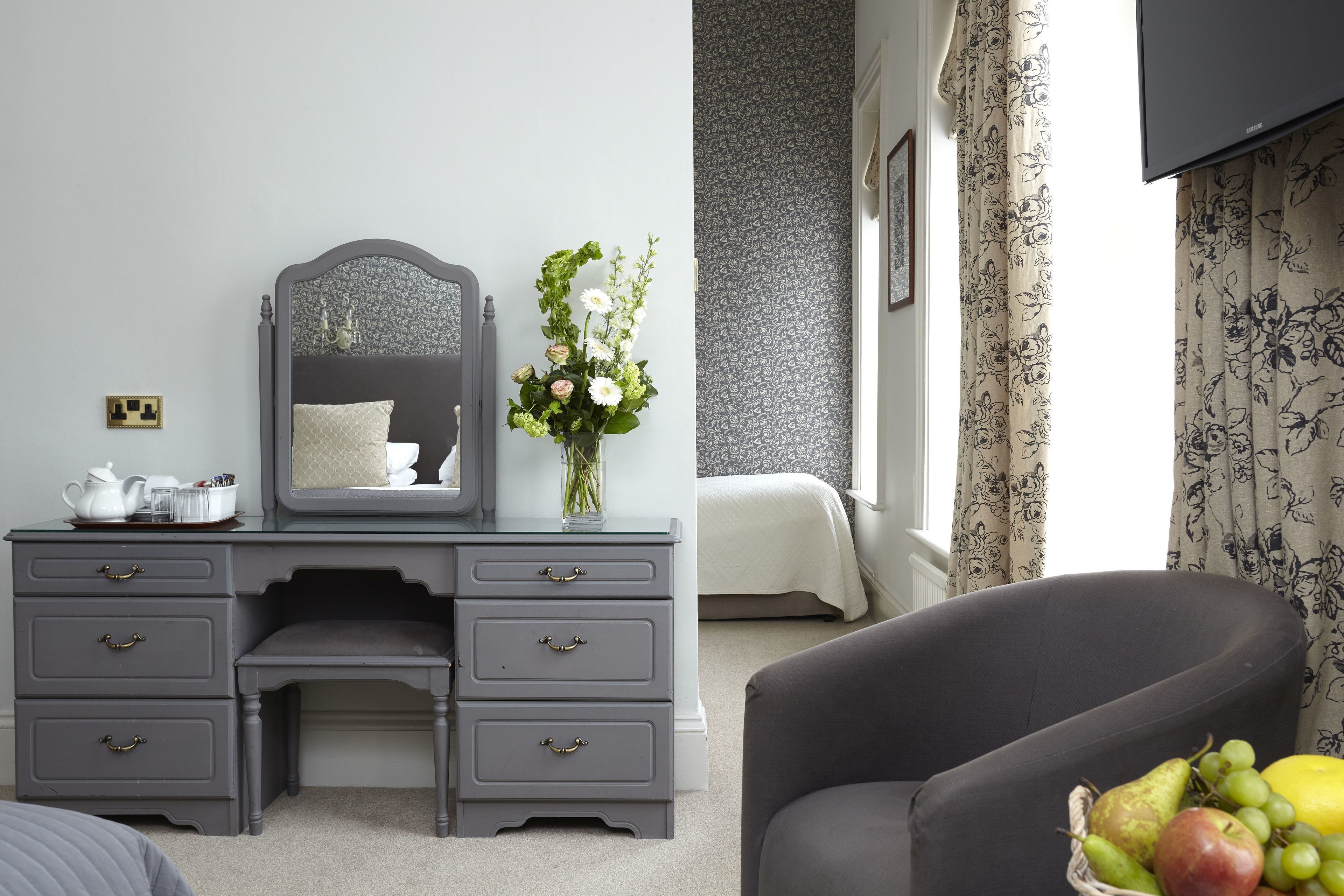 A bedroom with a grey dresser, a mirror, a vase with flowers, a bed with white bedding, and a chair with a fruit basket.