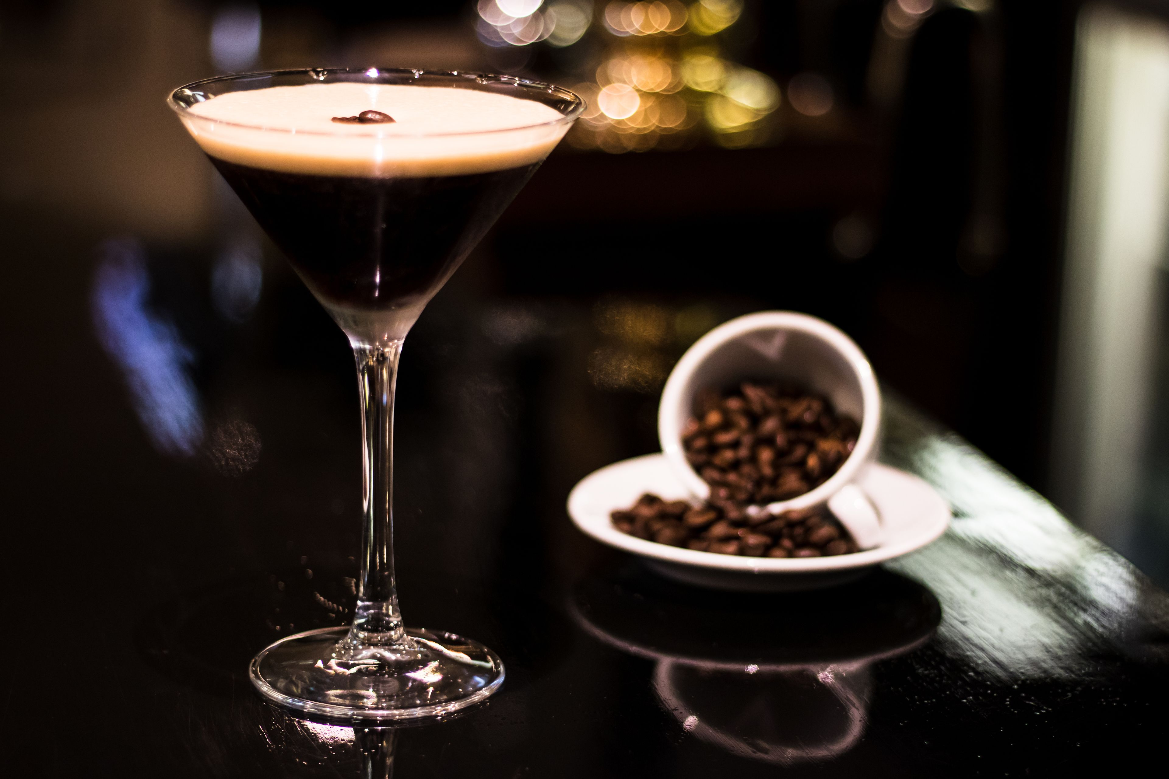 A martini glass with a dark liquid and foam on a reflective surface, a small bowl of coffee beans, and a blurred background with lights.