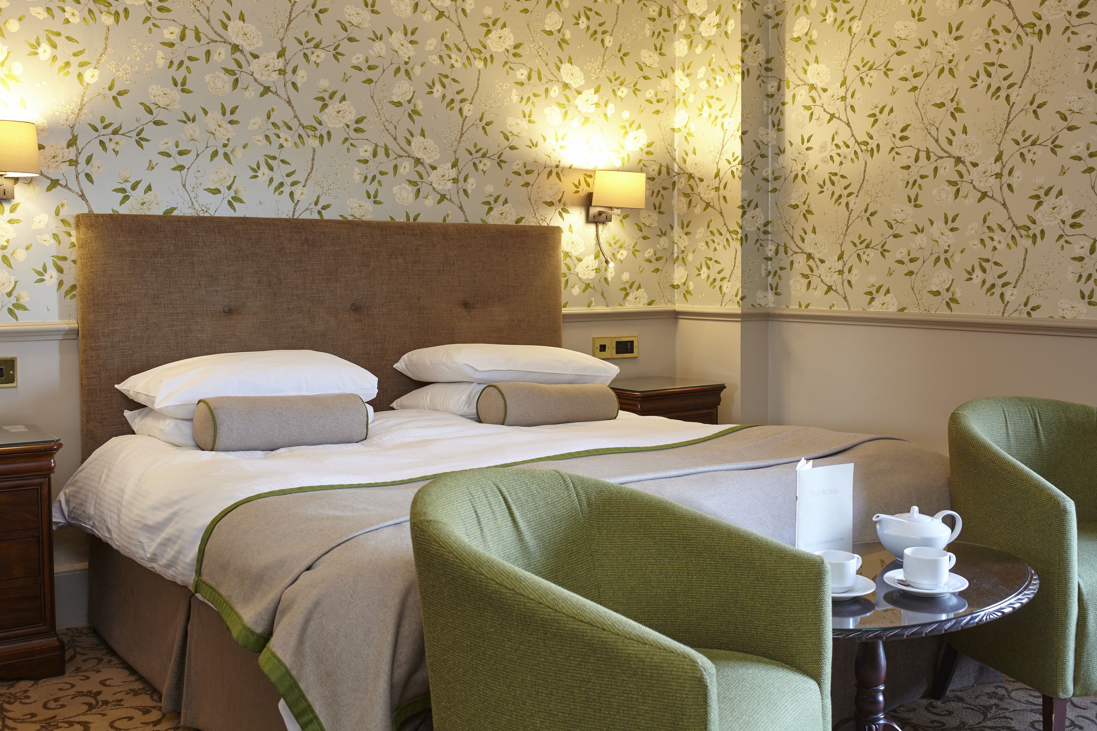 Habitación de hotel con una cama grande, sillones tapizados en verde, una mesita con tetera y tazas, y papel tapiz con motivos florales.