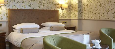 Habitación de hotel con una cama grande, sillones tapizados en verde, una mesita con tetera y tazas, y papel tapiz con motivos florales.