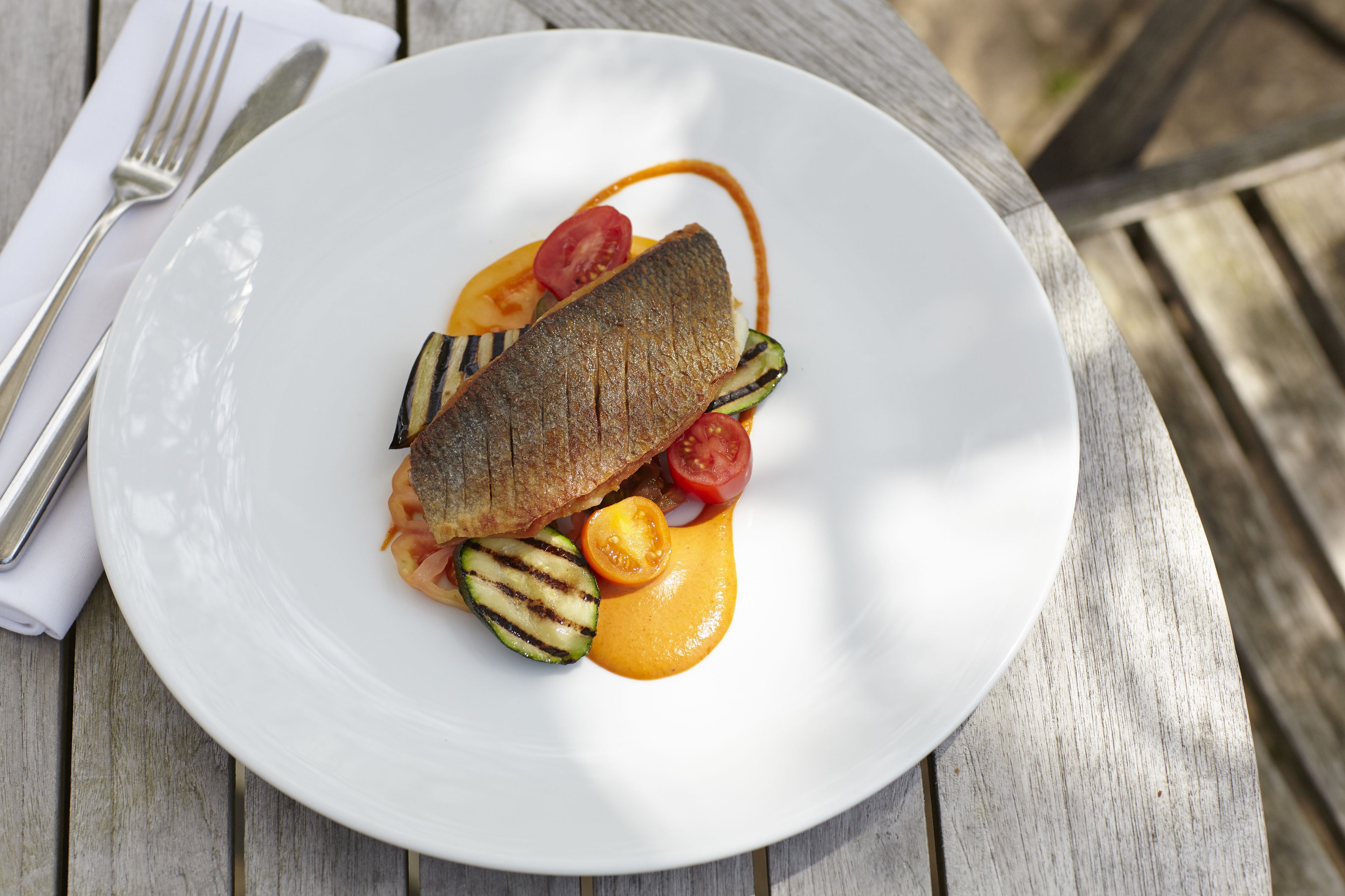 A white plate with grilled fish, cherry tomatoes, zucchini, and a sauce on a wooden table.