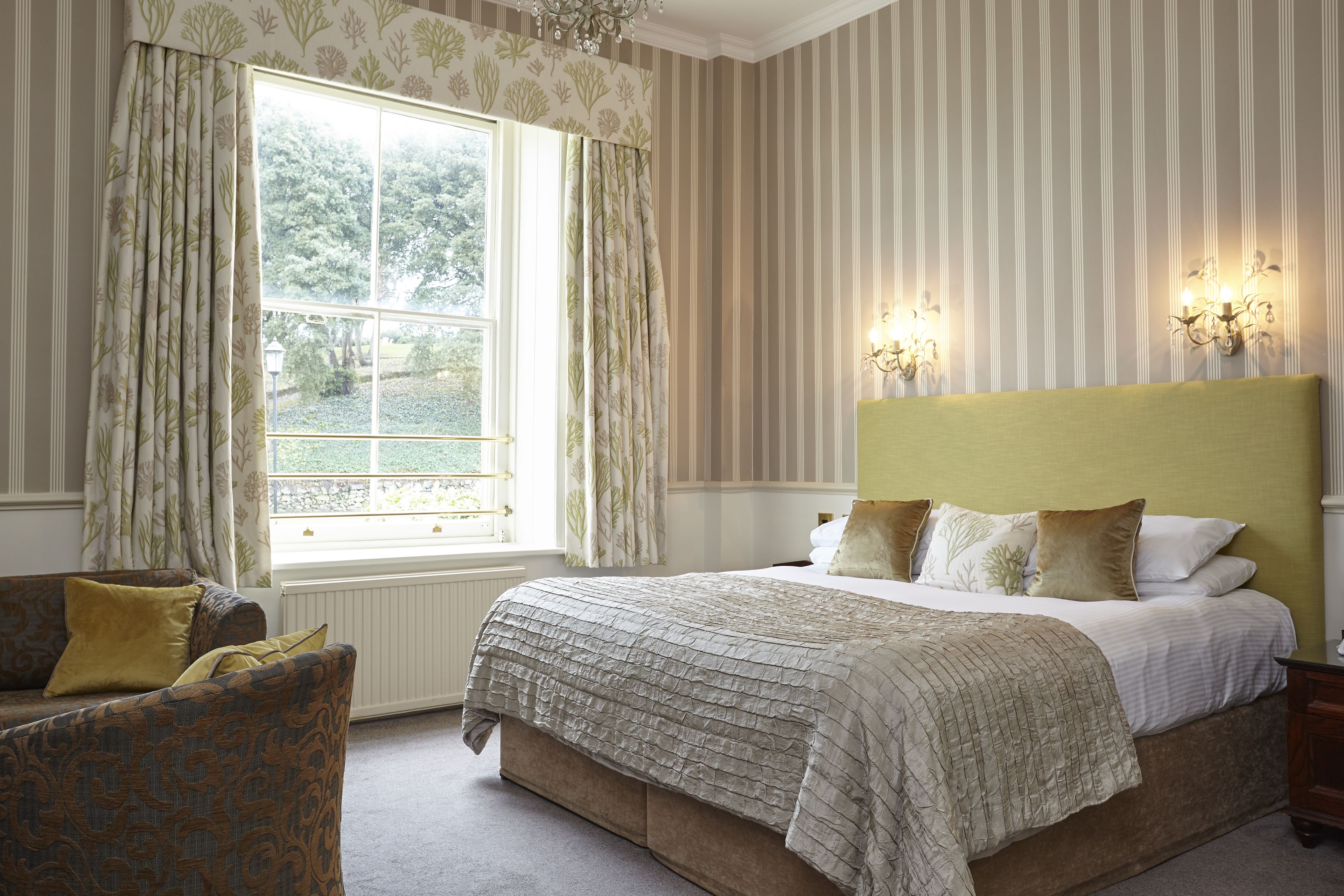 A bedroom with a large bed, a window with curtains, a chair, and wall sconces.